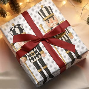 Op de afbeelding: Een ingepakt cadeau met een rode strik en een notenkrakerontwerp. Het cadeaupapier is wit met kleine grijze stippen. Het cadeau is omgeven door kerstversieringen en dennentakken.