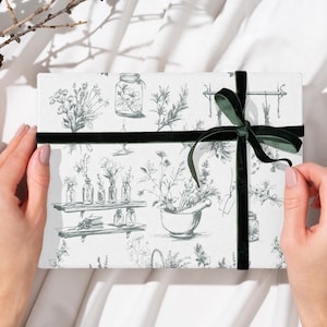 Puede incluir: Una caja de regalo rectangular envuelta en papel blanco con una ilustración botánica en tonos grises. La caja está atada con una cinta de terciopelo verde oscuro. Una ramita de piñas y ramas está en el fondo.