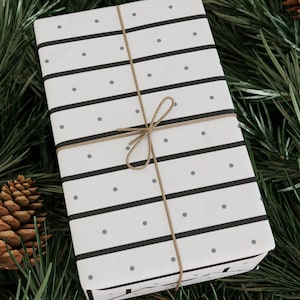May include: A white gift box with black stripes and gray polka dots wrapped with brown twine. The box is sitting on a bed of green pine needles.