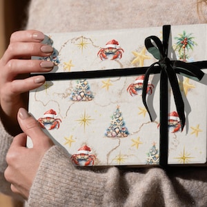 May include: A white gift wrap with a festive Christmas design featuring red crabs wearing Santa hats, Christmas trees decorated with seashells, and gold stars. The wrapping paper is tied with a black velvet ribbon.