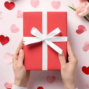 May include: A red gift box with a white ribbon and bow. The box is being held by a person's hands. The background is pink with hearts.