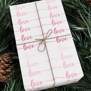 Op de afbeelding: Wit cadeaupapier met een herhalend patroon van het woord "love" geschreven in roze cursief. Het cadeau is vastgebonden met een bruin touwtje.