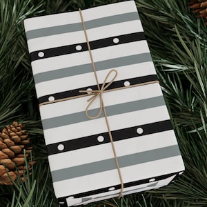 May include: A white gift box with black and gray stripes and white polka dots. The box is tied with a brown twine bow and is sitting on a bed of green pine needles.