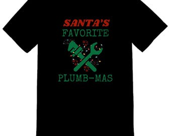 Pode incluir: Camiseta preta com o texto "SANTA'S FAVORITE PLUMB-MAS" em vermelho e verde. O design apresenta chaves inglesas verdes cruzadas e enfeites natalinos festivos, ideal para encanadores.