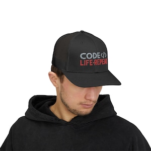 May include: Black trucker hat featuring the text "CODE<>LIFE-REPEAT" in silver and red. The hat has a mesh back and a curved peak. The hat is worn by a person wearing a black hoodie.