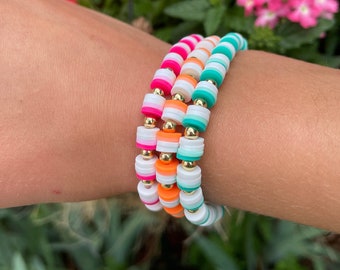 May include: Three colorful beaded bracelets stacked on a wrist. The bracelets are made of white, orange, pink, and teal beads with gold accents.