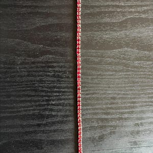 May include: A silver bracelet with a line of small, vibrant red gemstones. The bracelet is thin and delicate, with a simple clasp. The gemstones are evenly spaced and create a continuous line of color. The bracelet is displayed on a dark wooden surface.