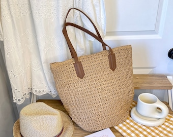 May include: A woven straw tote bag with brown leather handles and accents. A straw hat with a red and blue band sits next to the bag. A small book with the text "She BELIEVED She COULD so SHE DID" is also visible.