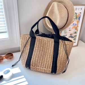 May include: A woven straw tote bag with black handles and accents. The bag has a drawstring closure. A straw hat and a framed print are in the background. The bag is on a white surface.