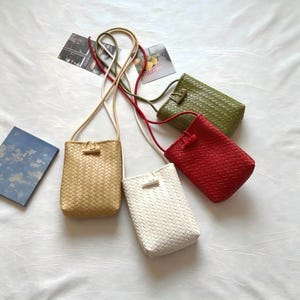 May include: Four rectangular crossbody bags with a woven texture, each featuring a long shoulder strap and a small toggle closure. The bags are available in beige, white, red, and olive green, each with a matching strap and a simple toggle fastener.