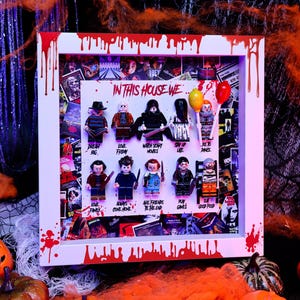 Könnte beinhalten: Ein weißer Schattenfugenrahmen mit roten Farbtropfen, der zehn Horrorfilm-Minifiguren in zwei Reihen enthält. Der Text "IN THIS HOUSE WE" ist oben aufgedruckt, mit individuellen Phrasen unter jeder Figur, wie "DREAM BIG" und "PLAY GAMES."