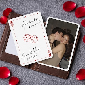 Può includere: Due carte da gioco, un portafoglio in pelle marrone e petali di rosa rossi sparsi. Una carta presenta il testo "How lucky are we?" e illustrazioni di dadi. L'altra carta mostra una foto di una coppia. È visibile anche il testo "Anna & Nick 04.11.26".