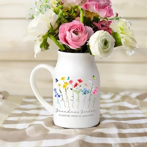 May include: White ceramic milk jug vase with a floral design and the text "Grandma's Garden Planting Seeds of Greatness". The vase has a handle and is decorated with colorful flowers and butterflies. Each flower has a name written on the stem.