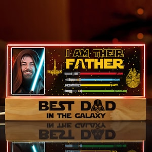 K&ouml;nnte beinhalten: Ein beleuchtetes Holzschild mit Star Wars-Motiv. Das Schild zeigt ein Bild von Obi-Wan Kenobi, ein Lichtschwert, einen Sternenzerst&ouml;rer und den Text "Ich bin ihr Vater." Unter dem Bild befindet sich eine Liste mit Namen: Liam, Noah, Lucy und Caroline. Am unteren Rand des Schildes steht "Bester Papa der Galaxie".