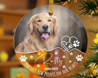 May include: A clear glass Christmas ornament with a photo of a golden retriever dog. The ornament has a brown background with white snowflakes, paw prints, and hearts. The text "Rusty 2006-2022 You left paw prints on our hearts" is printed on the ornament.