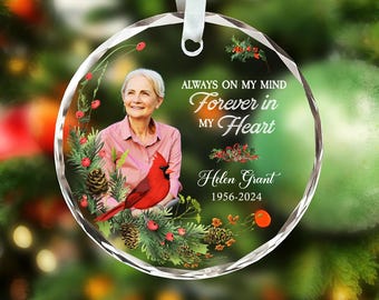 May include: A clear, round Christmas ornament with a photo of a woman and a red cardinal. Text reads "Always on my mind, Forever in my Heart, Helen Grant 1956-2024". The ornament has a white ribbon for hanging.
