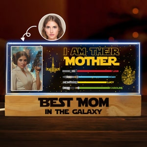 K&ouml;nnte beinhalten: Ein leuchtendes Schild im Star Wars-Stil mit einem Bild einer Frau in einem wei&szlig;en Outfit, die einen Blaster h&auml;lt. Das Schild lautet "Ich bin ihre Mutter" mit den Namen Liam, Noah, Lucy und Caroline darunter. Das Schild enth&auml;lt auch ein Bild eines Raumschiffs und Lichtschwerter. Die Basis des Schildes lautet "Beste Mama der Galaxie".