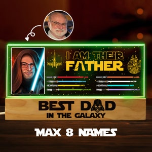 K&ouml;nnte beinhalten: Ein gr&uuml;n-schwarzes beleuchtetes Schild mit Star Wars-Thema. Das Schild zeigt ein Portr&auml;t eines Mannes in einem braunen Kapuzengewand, ein Lichtschwert, ein Raumschiff und den Text "Ich bin ihr Vater". Unter dem Portr&auml;t befinden sich acht Zeilen Text mit Namen, jede mit einer farbigen Lichtschwertgrafik. Das Schild zeigt auch den Text "Bester Papa der Galaxie" und eine Grafik von Darth Vader. Das Schild steht auf einem Holzsockel mit dem Text "Max 8 Namen".