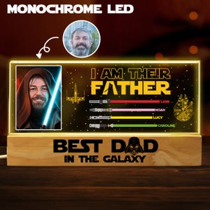 K&ouml;nnte beinhalten: Ein Holzsockel mit einer leuchtenden Acrylplatte mit einem Star Wars-Motiv. Die Platte zeigt eine Figur aus Star Wars, ein Raumschiff und Lichtschwerter. Der Text "Ich bin ihr Vater" und "Bester Papa der Galaxie" ist auf der Platte zu sehen. Ein Foto eines Mannes befindet sich in der oberen linken Ecke des Bildes.