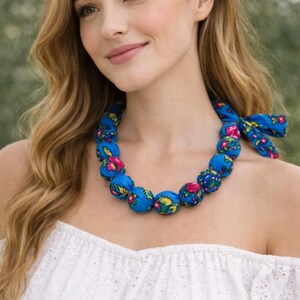Puede incluir: Un collar de cuentas de tela azul con un estampado floral en rosa, amarillo y verde. El collar está atado con un lazo de tela a juego. La mujer lleva un top blanco con los hombros descubiertos.