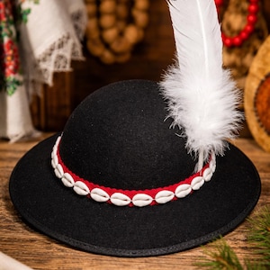 May include: A black hat with a red band adorned with white seashells. A large white feather with a black tip is attached to the hat. The hat is displayed on a wooden surface, with floral textiles in the background.