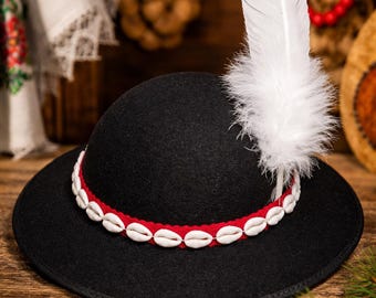 May include: A black hat with a red band adorned with white seashells. A large white feather with a black tip is attached to the hat. The hat is displayed on a wooden surface, with floral textiles in the background.