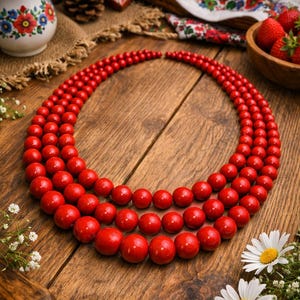 May include: A three-strand red beaded necklace. The necklace is composed of round, glossy red beads of varying sizes. The necklace is displayed on a wooden surface, with daisies and other decorative elements in the background.