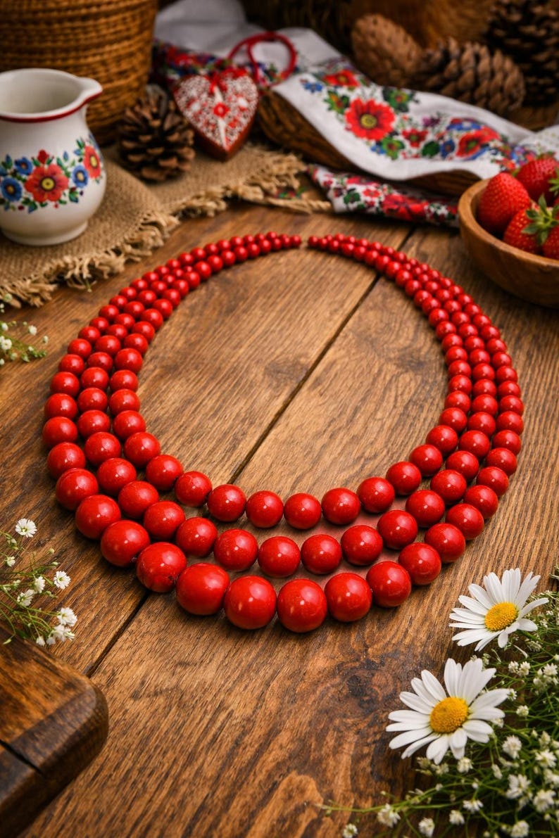 May include: A three-strand red beaded necklace. The necklace is composed of round, glossy red beads of varying sizes. The necklace is displayed on a wooden surface, with daisies and other decorative elements in the background.