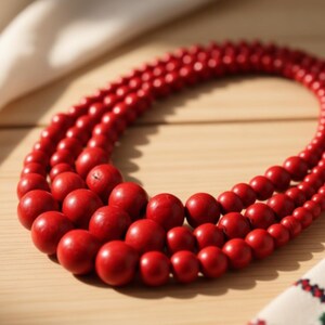 May include: A three-strand necklace featuring vibrant red wooden beads. The beads vary in size, creating a layered effect. The necklace is displayed on a light wooden surface, with a white cloth and a patterned textile in the background.