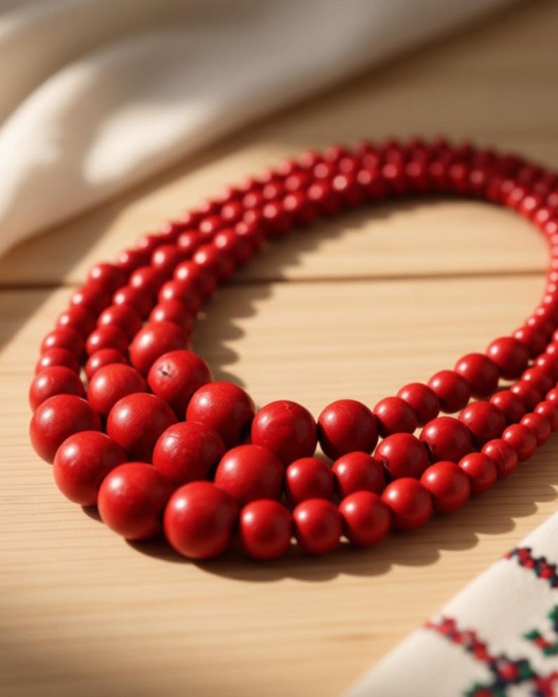 May include: A three-strand necklace featuring vibrant red wooden beads. The beads vary in size, creating a layered effect. The necklace is displayed on a light wooden surface, with a white cloth and a patterned textile in the background.