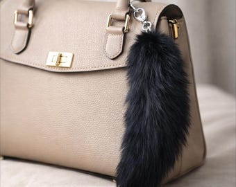 May include: A beige leather handbag with gold-tone hardware and a black fur keychain attached. The handbag has a structured design with a top handle and a flap closure. The fur keychain is long and fluffy.