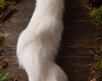 May include: A fluffy, white faux fur tail with a silver-colored ring at the top. The tail is curved and has a soft, textured appearance. The background includes moss, pinecones, mushrooms, and a lit candle, suggesting a woodland theme.