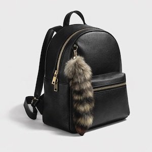 Puede incluir: Mochila negra con cremalleras y herrajes dorados. La mochila tiene un pequeño bolsillo frontal y un asa superior. Un adorno de cola de piel sintética esponjosa cuelga de la cremallera. La mochila está hecha de un material liso.