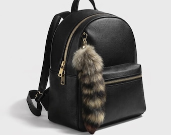 May include: Black backpack with gold-colored zippers and hardware. The backpack has a small front pocket and a top handle. A fluffy, faux fur tail charm hangs from the zipper. The backpack is made of a smooth material.