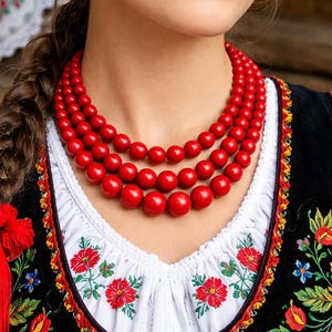 May include: A woman wearing a traditional Polish outfit, including a white blouse with red floral embroidery and a black vest. A triple-strand red bead necklace is the focal point. The outfit is a vibrant display of Polish folk culture.