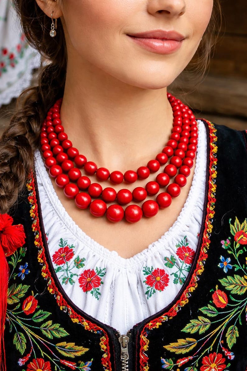 May include: A woman wearing a traditional Polish outfit, including a white blouse with red floral embroidery and a black vest. A triple-strand red bead necklace is the focal point. The outfit is a vibrant display of Polish folk culture.