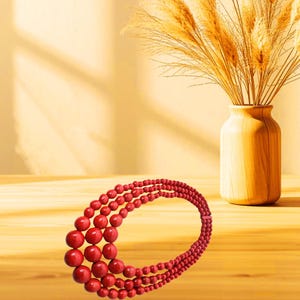 May include: A multi-strand red beaded necklace. The necklace features large, round beads that gradually decrease in size. The necklace is displayed on a wooden surface, with a wooden vase and dried wheat stalks in the background.