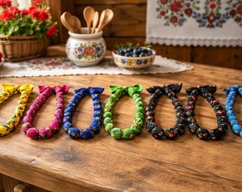 May include: A collection of colorful, fabric-covered necklaces in yellow, pink, blue, green, black, and blue. Each necklace is adorned with a bow. The necklaces are displayed on a wooden table, with a rustic, floral-themed background.