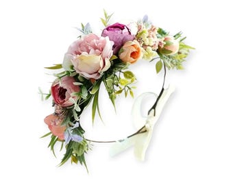 May include: A floral crown featuring a variety of artificial flowers in shades of pink, peach, and purple, accented with green leaves. The flowers are arranged on a thin, circular wire base with a cream-colored ribbon.