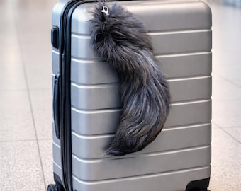 May include: A silver rolling suitcase with a black handle and wheels. A fluffy, gray tail charm hangs from the handle. The suitcase has a ribbed design and is in an airport setting.
