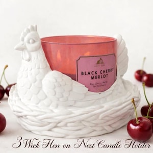 May include: A white ceramic hen-shaped candle holder with a pink candle labeled "Black Cherry Merlot." The holder is surrounded by fresh cherries. The text "3 Wick Hen on Nest Candle Holder" is at the bottom.