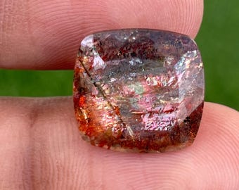 May include: A square-cut gemstone displaying a blend of red, orange, and brown hues. The translucent gem features internal patterns and inclusions. The stone is held between two fingers, highlighting its size and intricate details.