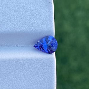 Op de afbeelding: Een peervormige, gefacetteerde edelsteen met een diepblauwe kleur. De edelsteen ligt op een wit, getextureerd oppervlak, met een vage groene achtergrond. De facetten van de edelsteen reflecteren licht, waardoor een sprankelend effect ontstaat.