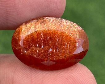 May include: A faceted, oval gemstone in shades of reddish-orange. The gem is held between fingers, with a blurred green background. The surface of the gem is reflective and sparkling. The gem is likely a sunstone.