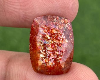 May include: A faceted, rectangular gemstone with a warm, reddish-brown base colour. The stone is filled with sparkling inclusions of gold, pink, and iridescent colours, creating a shimmering effect. The gem is held between two fingers against a blurred green background.