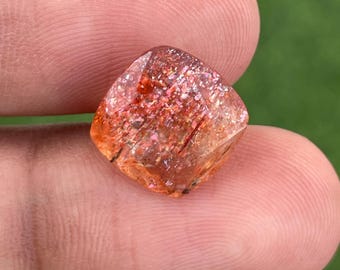May include: A square-cut sunstone gemstone with a sparkling, reddish-orange interior. The gem has a translucent appearance with visible inclusions and a polished surface. The stone is held between two fingers against a blurred green background.