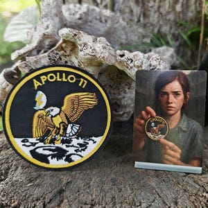 Peut inclure: Un écusson noir et jaune Apollo 11 avec un aigle et le texte "Apollo 11". Une petite carte montre une personne tenant un écusson similaire. Les articles sont exposés sur une surface en bois.