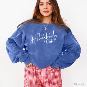 May include: A blue sweatshirt with the text "The Homebody Club" in white script. The sweatshirt has long sleeves and a crew neck. The model is wearing red and white checkered pajama pants. The color of the sweatshirt is "Flo Blue".