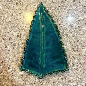 May include: A glossy, deep teal ceramic tray shaped like a pointed leaf. The tray features a raised central vein running vertically and a distinct, wavy, scalloped edge that follows the entire perimeter of the leaf-shaped form.