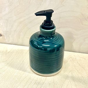 Pu&ograve; includere: Un dispenser di sapone cilindrico in ceramica con una smaltatura color verde acqua profondo e texture a coste orizzontali. La base &egrave; in ceramica bianca non smaltata e la parte superiore &egrave; dotata di un meccanismo a pompa in plastica nera.